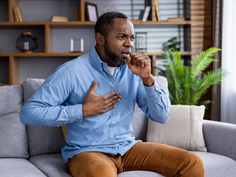 The image depicts a middle-aged male sitting in his chair, coughing, in discomfort, and clutching chest.