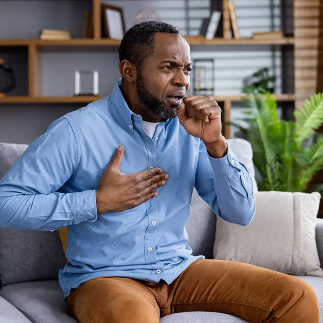 The image depicts a middle-aged male sitting in his chair, coughing, in discomfort, and clutching chest.