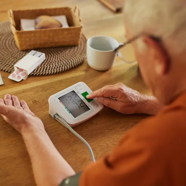 The image depicts an elderly man sits at the table with his pills set out in front of him. He also has a blood pressure machine and is testing his pressure.