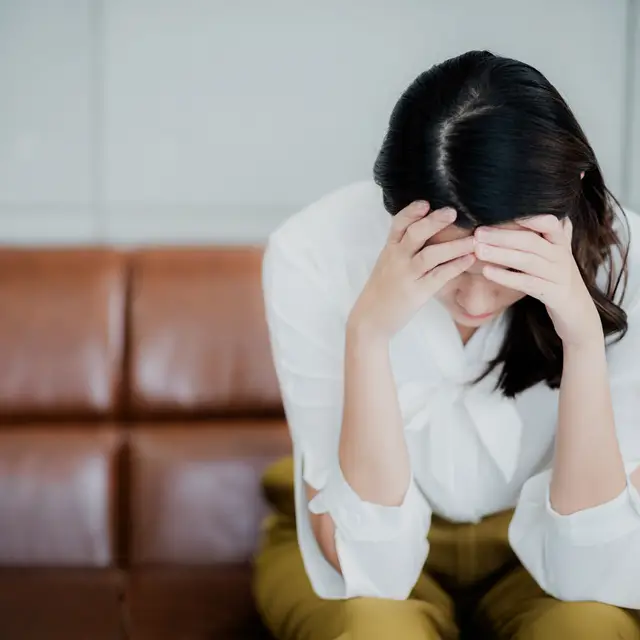 The image depicts a frustrated woman sits on a couch, hunched over with her face in her hands, seemingly stressed.